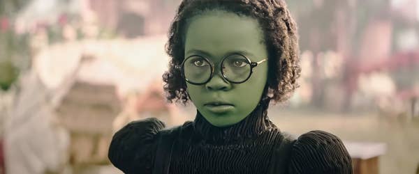 A person with green skin and round glasses wears a vintage-style black dress, appearing in a dramatic or fantasy setting
