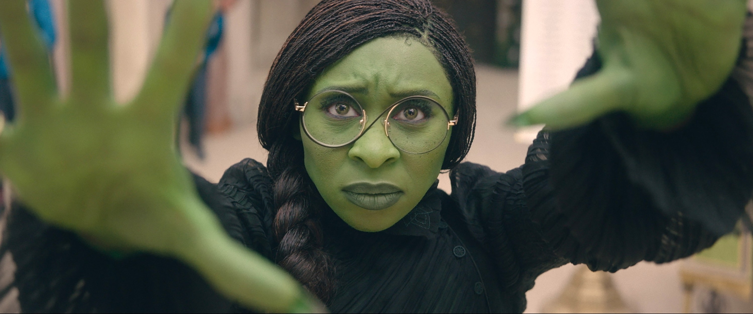 Person with green makeup and glasses, wearing a pleated outfit, extends hands towards the camera in a dramatic pose