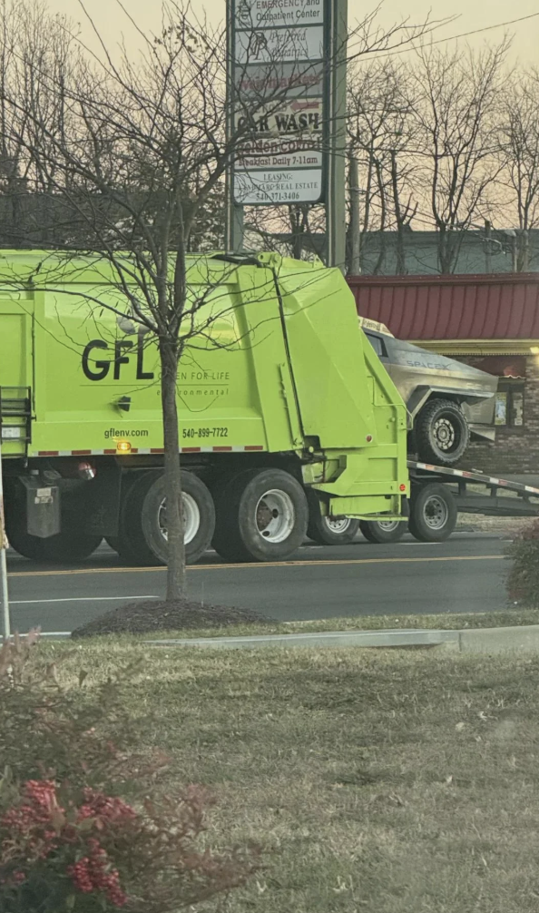 Garbage truck towing a vintage car on a trailer in a street setting with bare trees and background signage