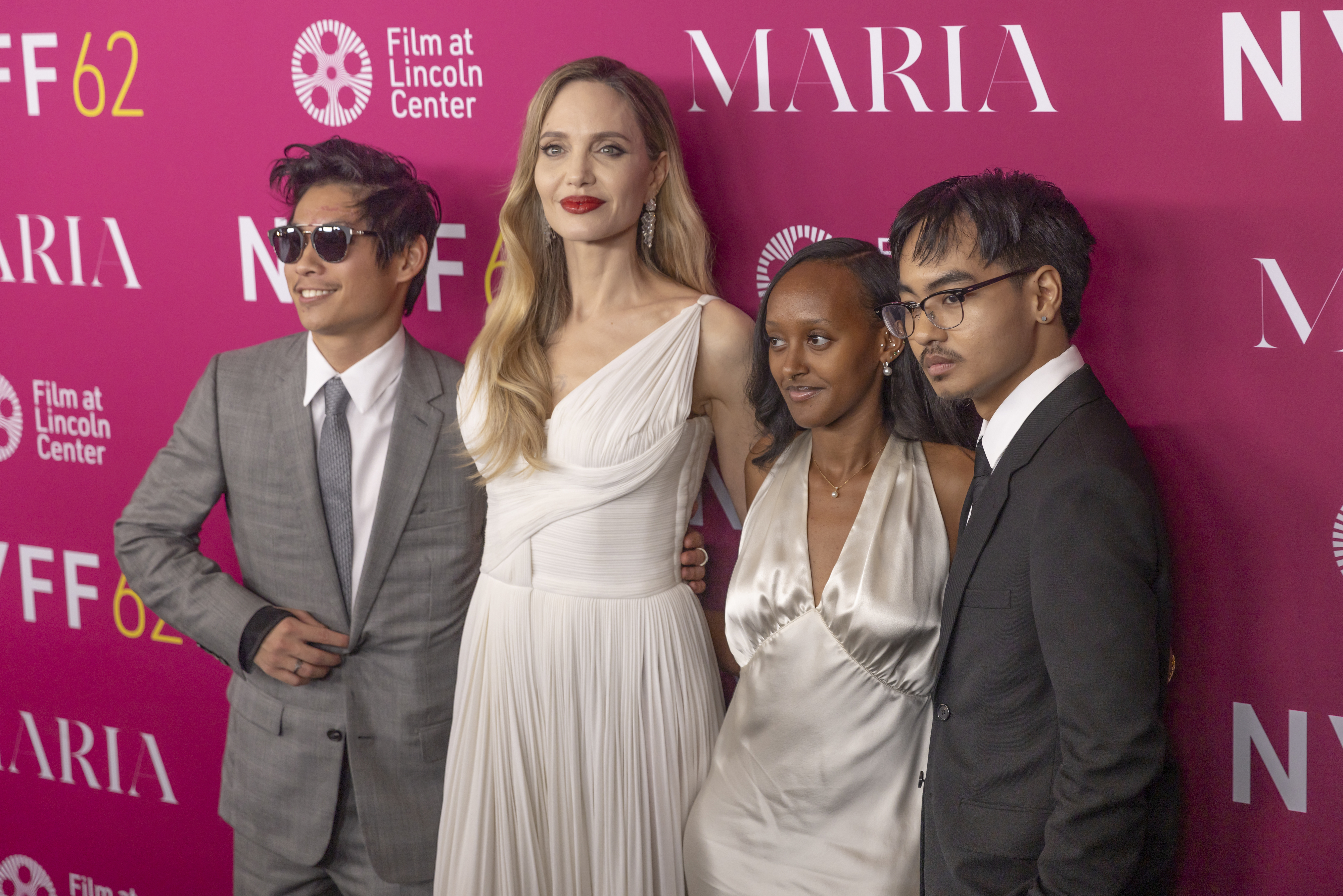Four people pose on a red carpet; one woman in an elegant white gown, and three men in suits