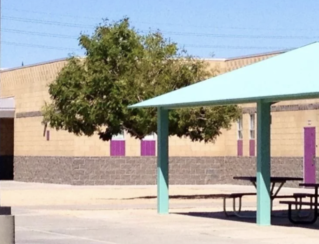 A school courtyard with a picnic table under a teal shade structure, and a tree near the building