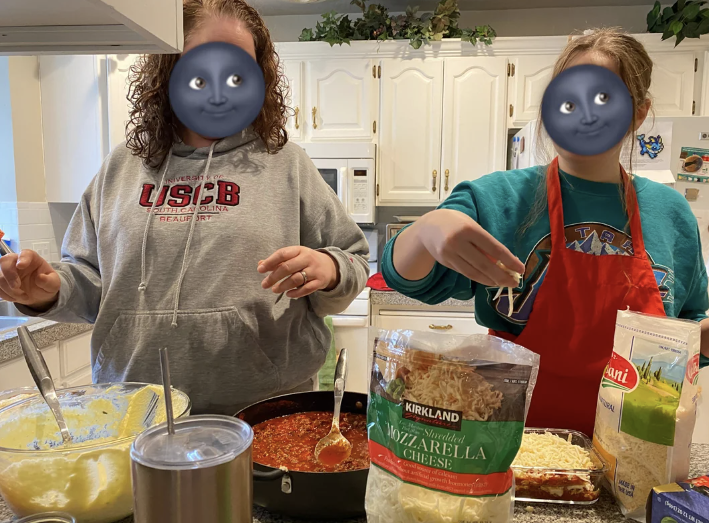 Two people cooking in a kitchen, preparing dishes with cheese and sauce. One wears a "USCB" hoodie; the other, a green shirt and red apron
