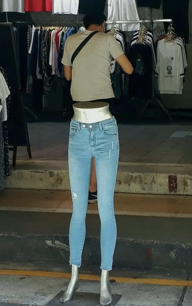Store display of headless mannequin in jeans, silver shoes; person in background adjusting t-shirts