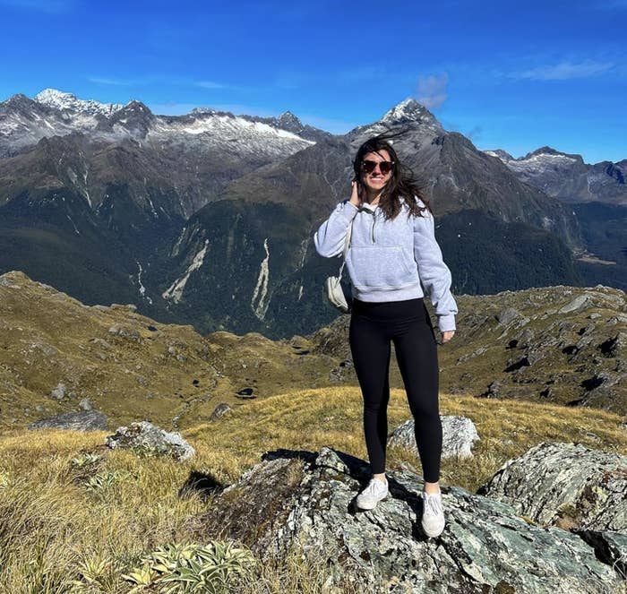 Person wearing a hoodie and leggings standing on a rocky hilltop with mountains in the background