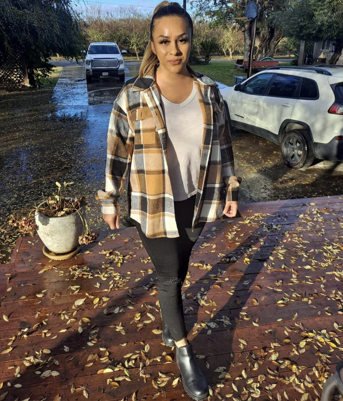 Person outdoors in autumn setting, wearing a plaid jacket, a light top, black pants, and ankle boots, walking on a leaf-covered pathway