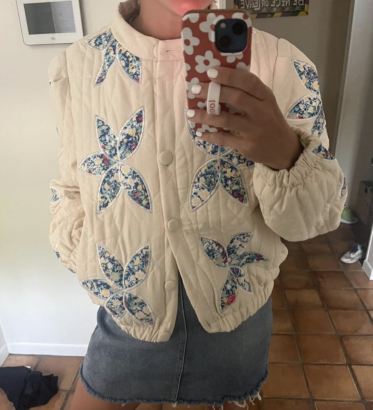 Person wearing a quilted jacket with floral patterns and a denim skirt, taking a mirror selfie