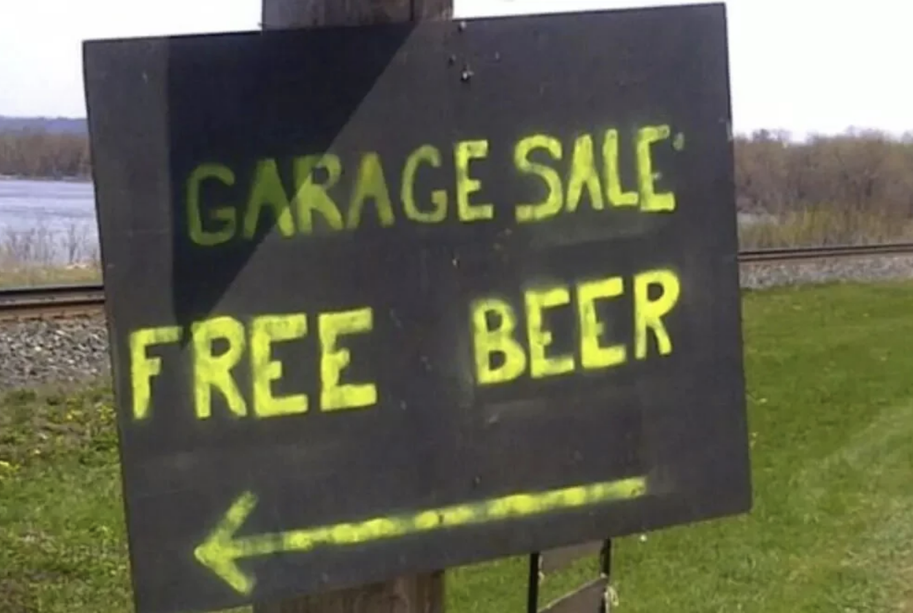Sign reading "Garage Sale Free Beer" with an arrow pointing left, set outdoors near a railway and grassy field
