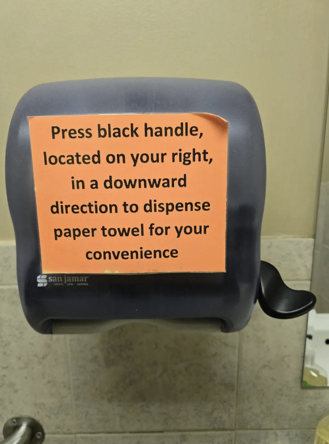 Paper towel dispenser with a label instructing to press the black handle on the right downward to dispense a towel