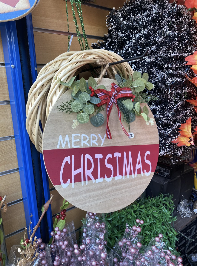 Wreath with wooden sign reading "Merry Chrismas" adorned with greenery and a red plaid bow, displayed in a store