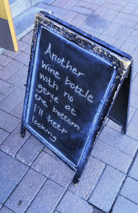 Chalkboard sign on sidewalk reads: "Another wine bottle with no genie at the bottom. I'll keep looking."