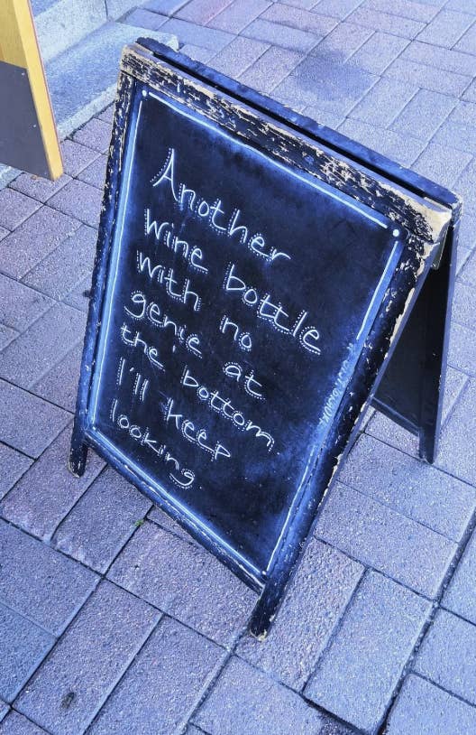 Chalkboard sign on sidewalk reads: &quot;Another wine bottle with no genie at the bottom. I&#x27;ll keep looking.&quot;