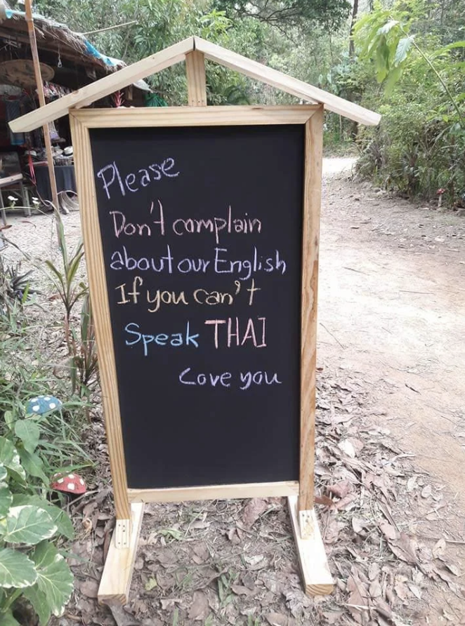 Chalkboard sign reads: "Please don't complain about our English if you can't speak Thai. Love you." Located outdoors on a path