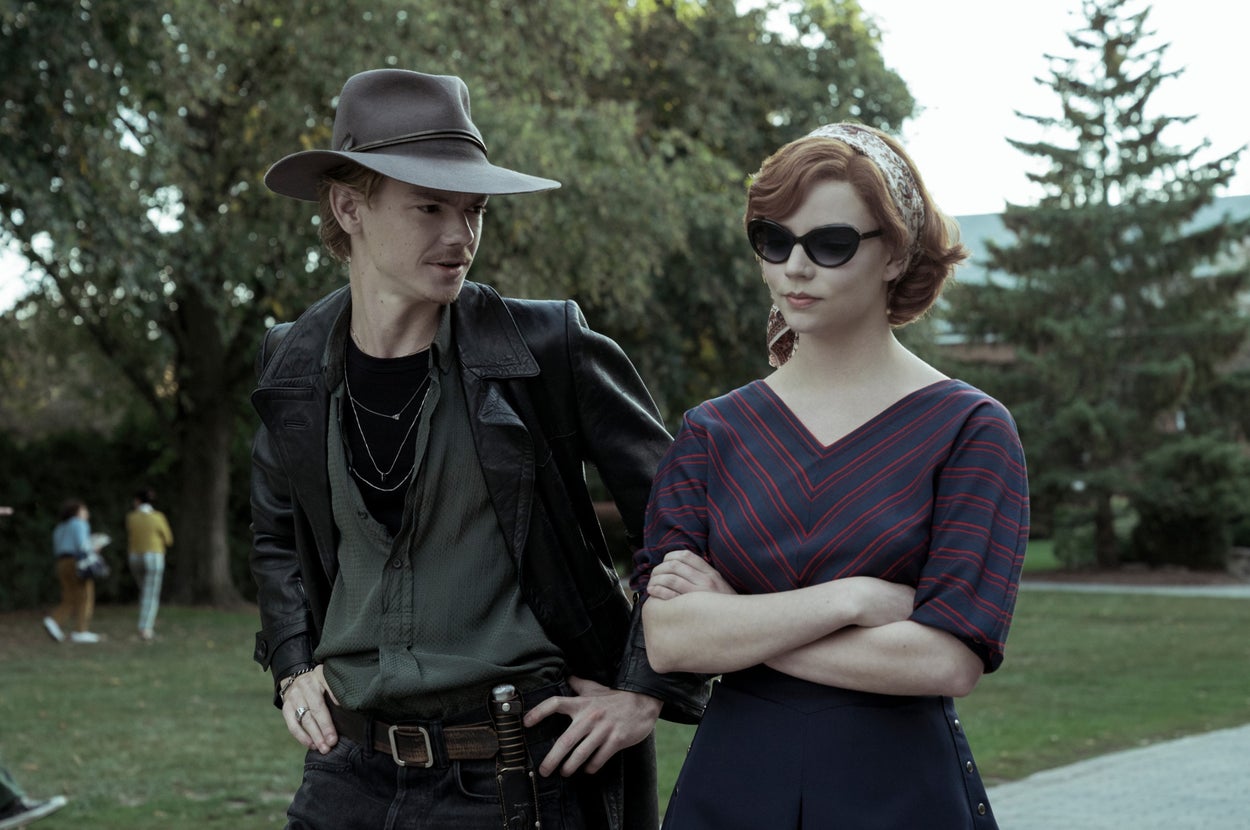 Two people outdoors from "Queen's Gambit," one wearing a cowboy hat and jacket with a gun holster, the other in a vintage-style dress with sunglasses and a headband