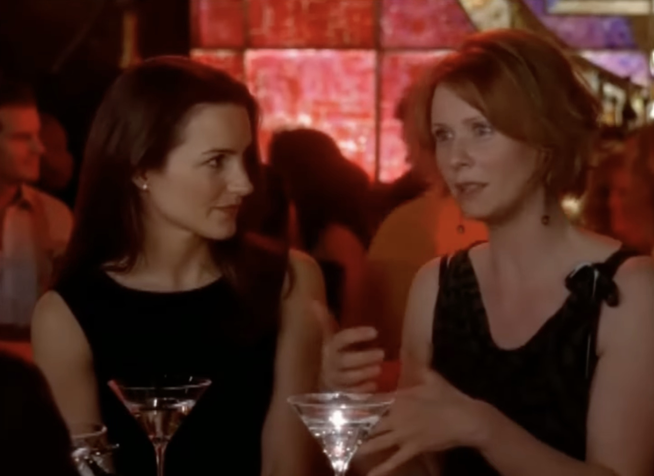 Two women talking at a bar, both holding martini glasses, surrounded by people and a colorful backdrop