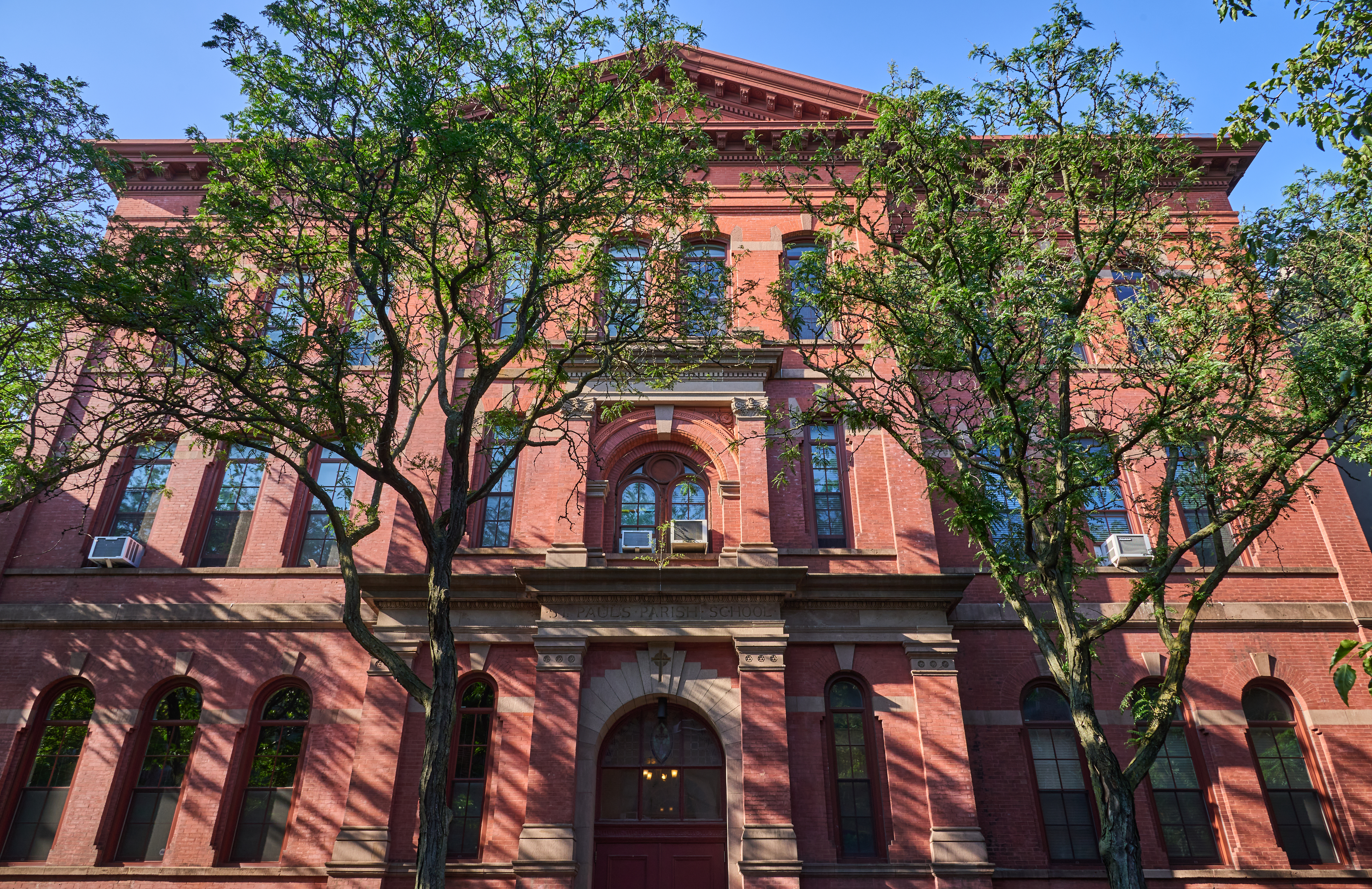 A historic brick building with arched windows and a central entrance, partially obscured by trees