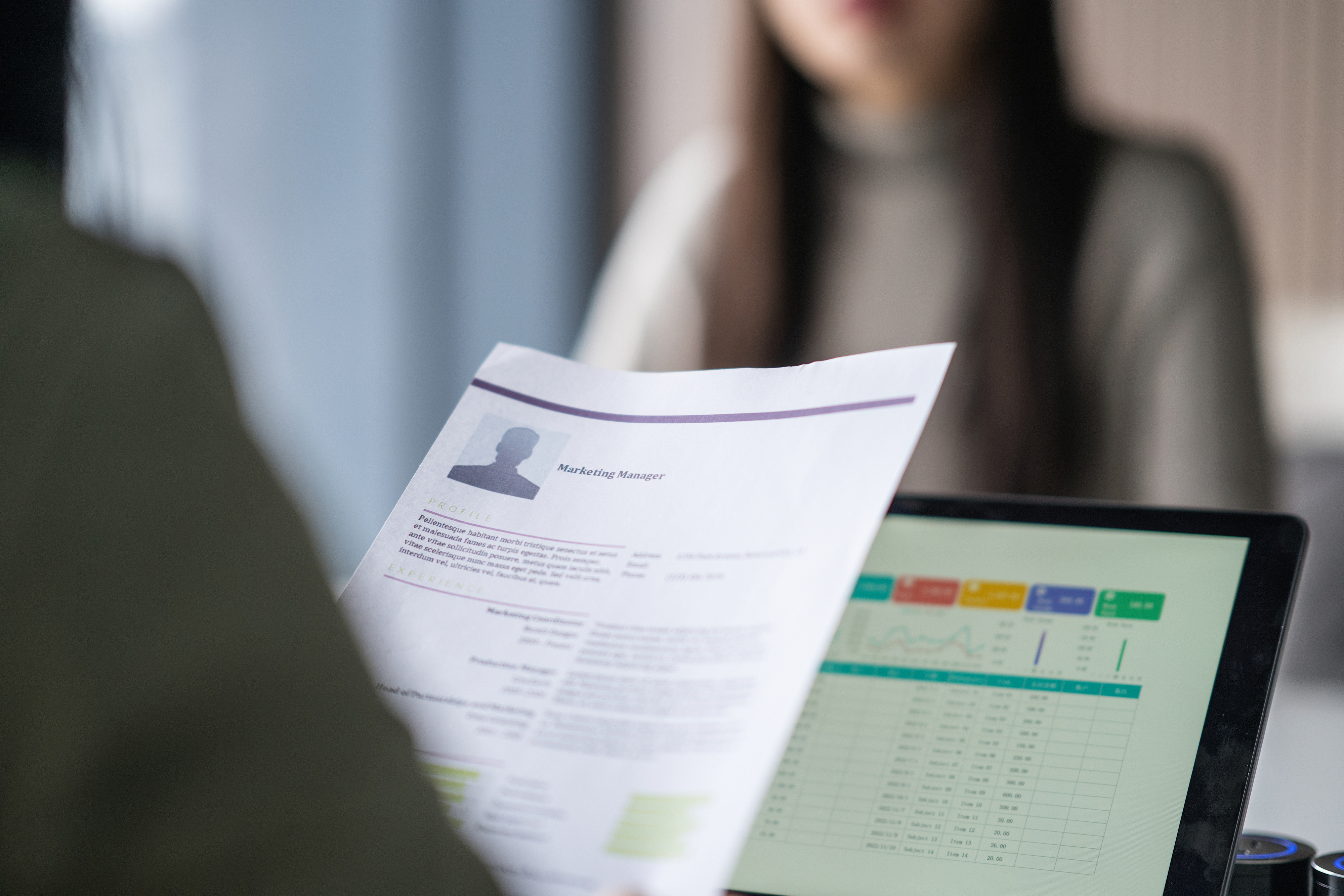 Person holding a printed resume titled &quot;Marketing Manager&quot; during an interview; a laptop with data charts is visible in the foreground