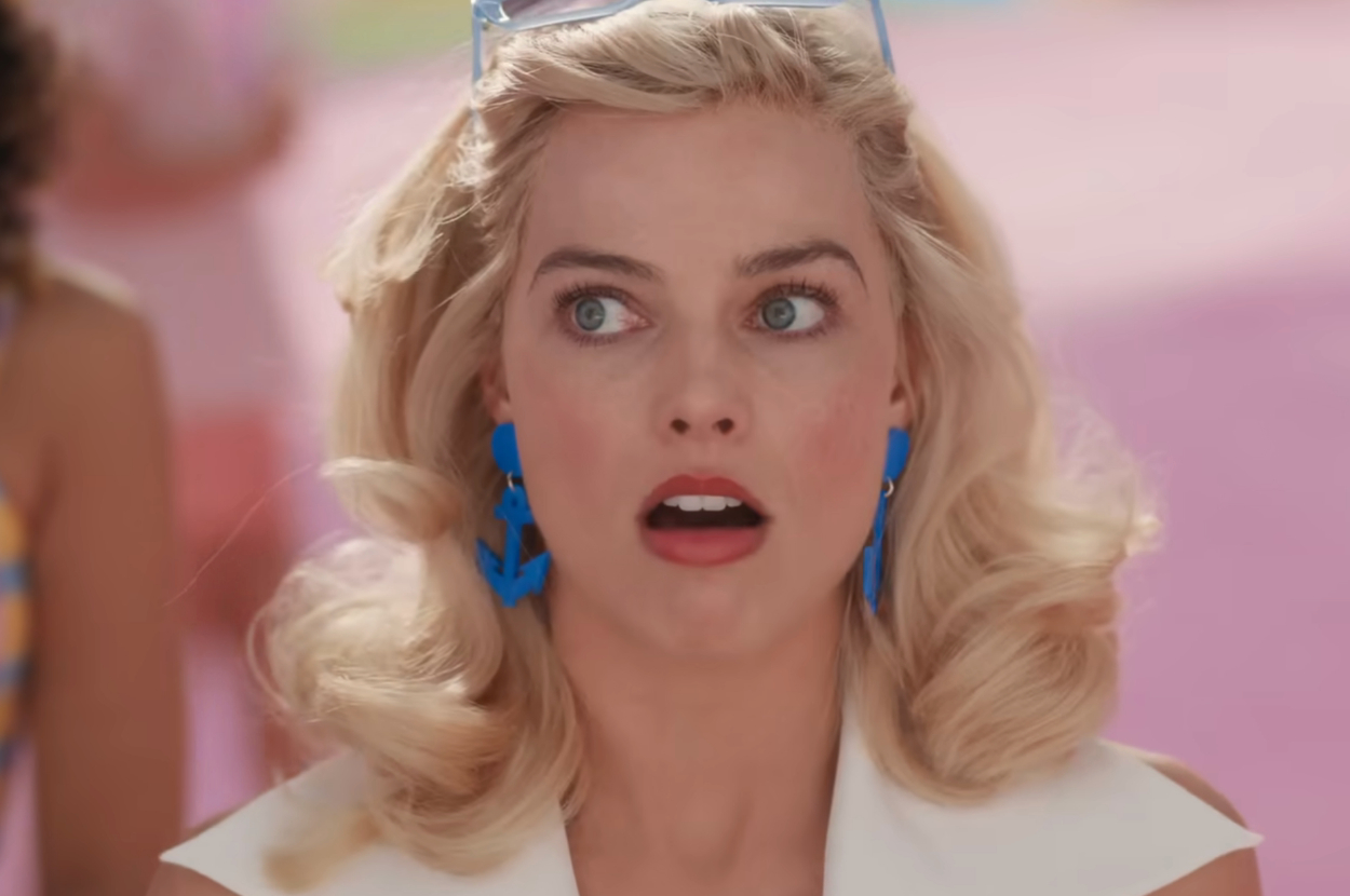 Actor looking surprised with styled blonde hair, wearing blue anchor earrings and a white outfit. Pastel background