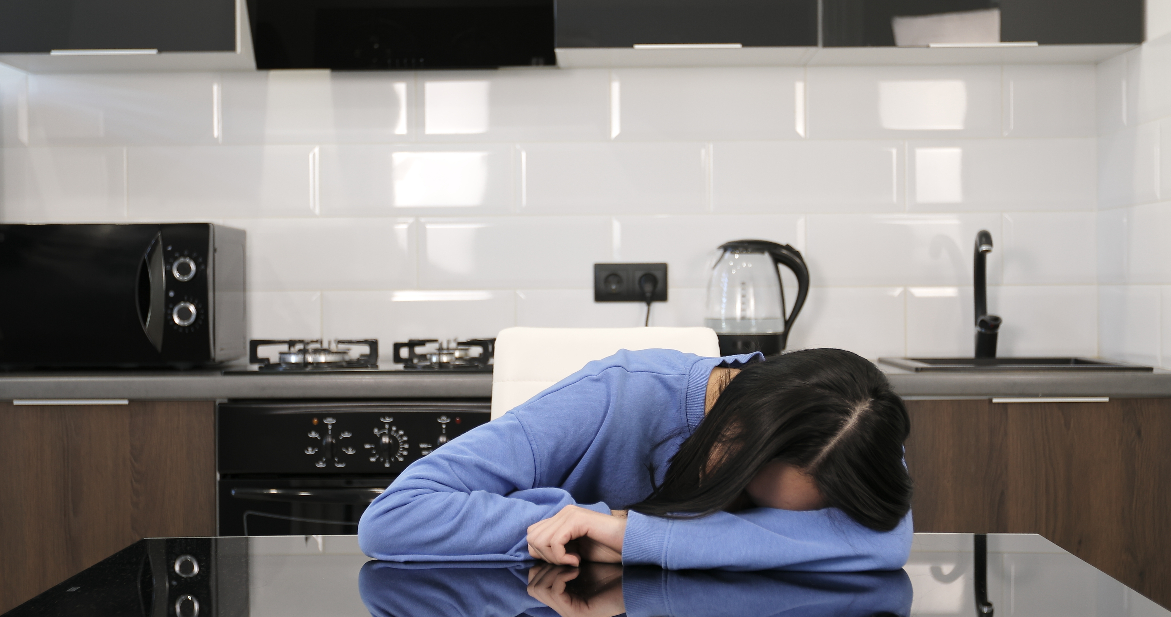 Person in a blue sweatshirt rests their head on their arms at a kitchen table with modern appliances in the background