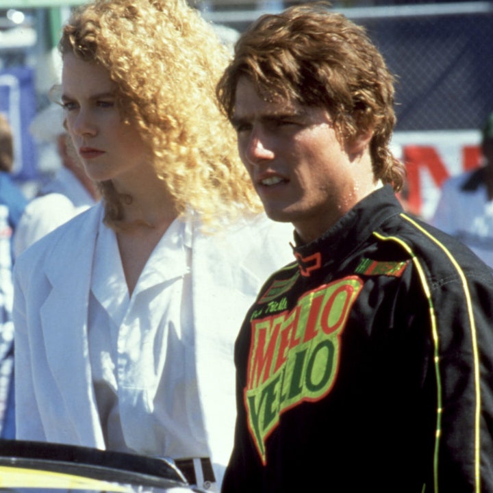 Two individuals at a racing event; one wears a racing suit with sponsor logos, the other in an elegant white outfit with curly hair
