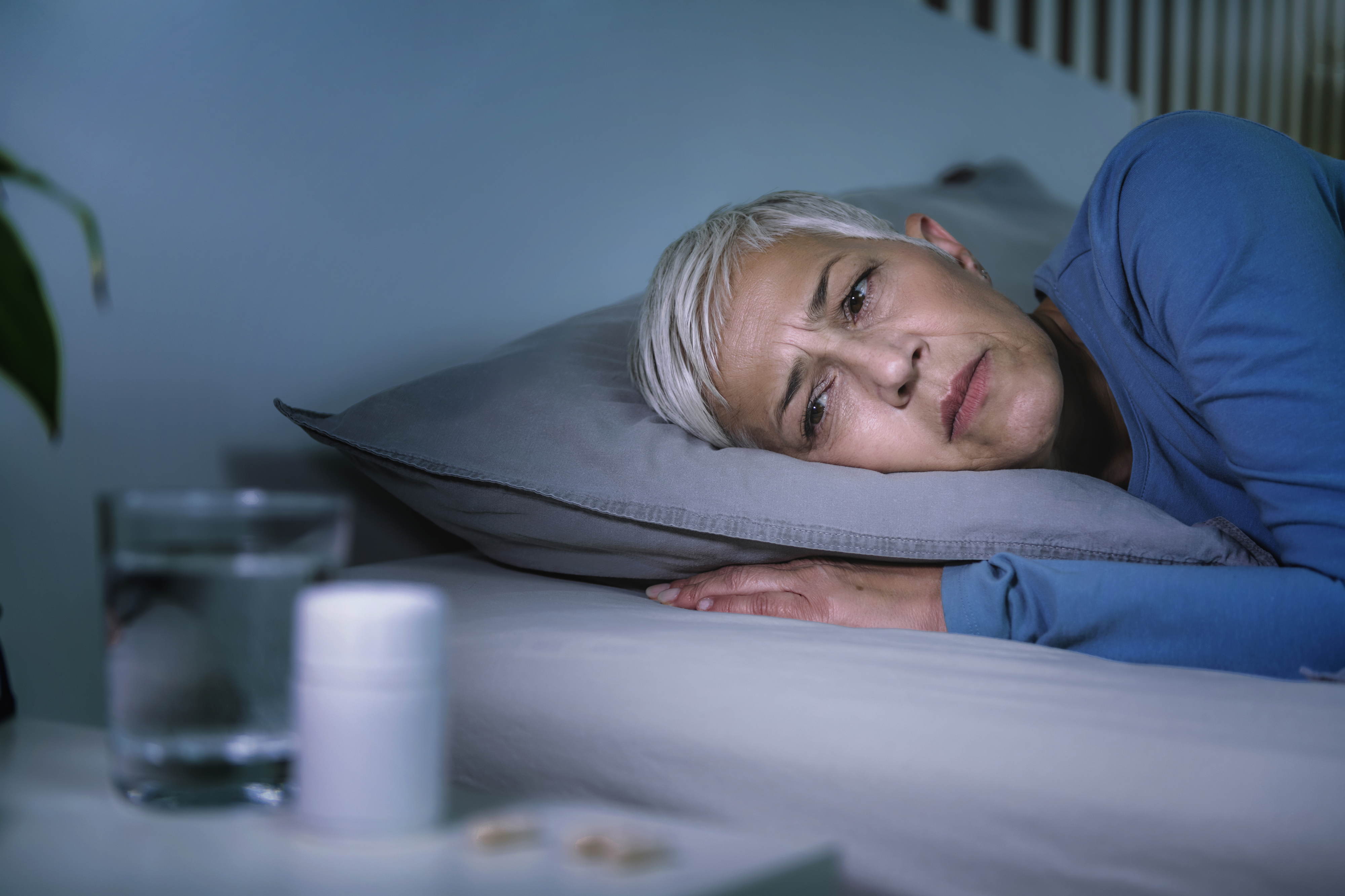 Person with short hair lies awake in bed, looking thoughtful. A glass of water and a pill bottle are on the bedside table