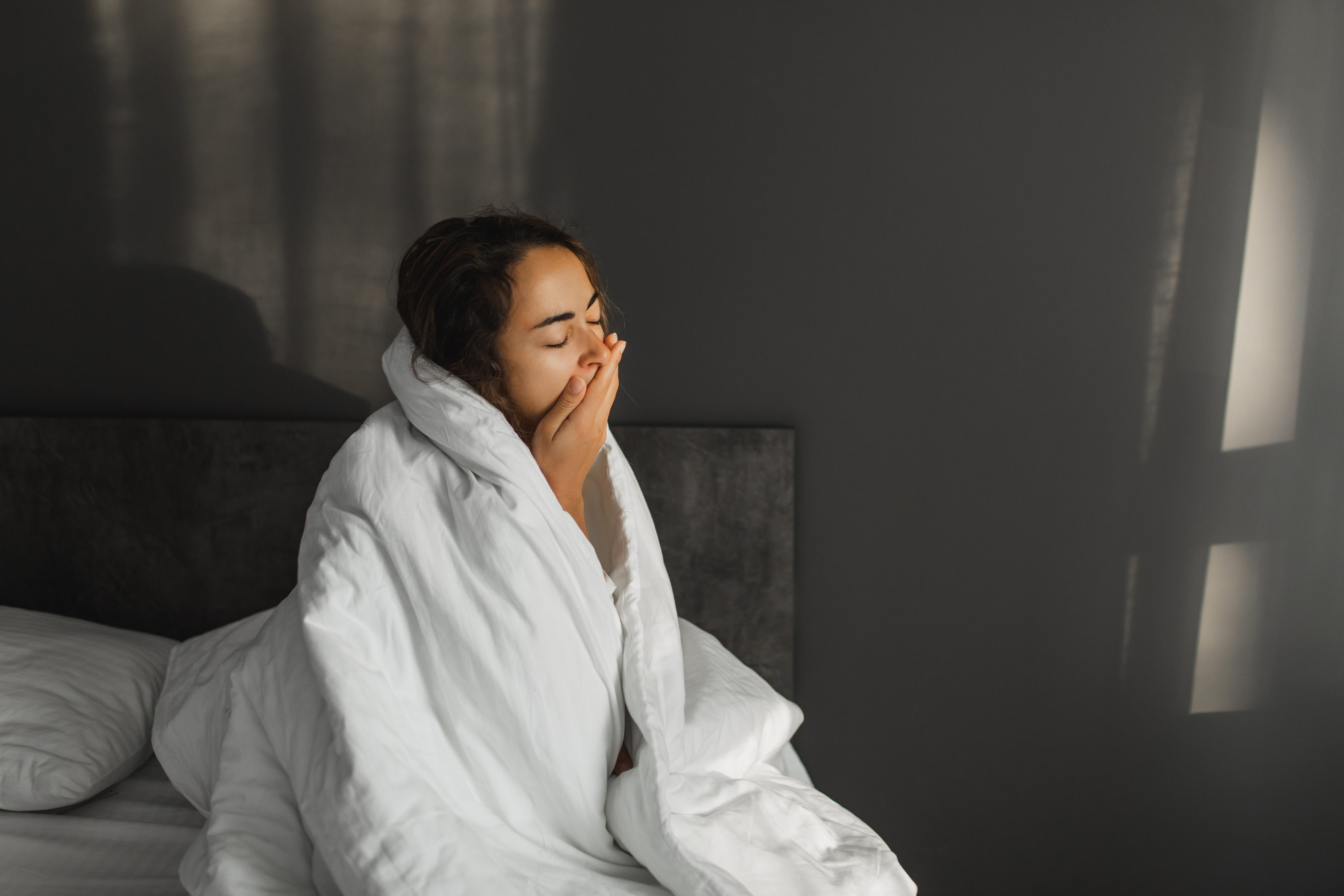 Person yawning in bed, wrapped in a white blanket, sitting up against a headboard in a softly lit room