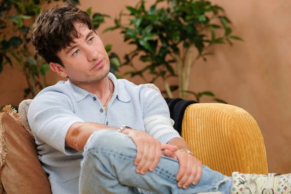 Barry Keoghan sitting on a couch wearing a sweater and jeans, appearing thoughtful, with plants in the background