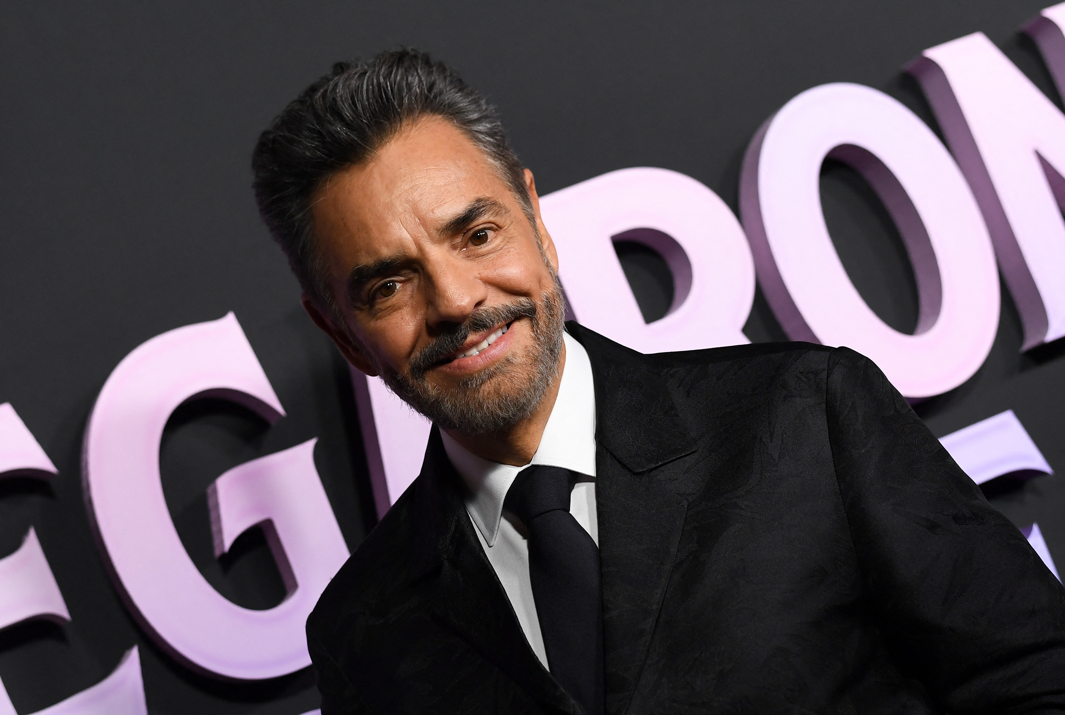 Eugenio Derbez in a sleek suit and tie at a celebrity event, smiling in front of a decorative backdrop