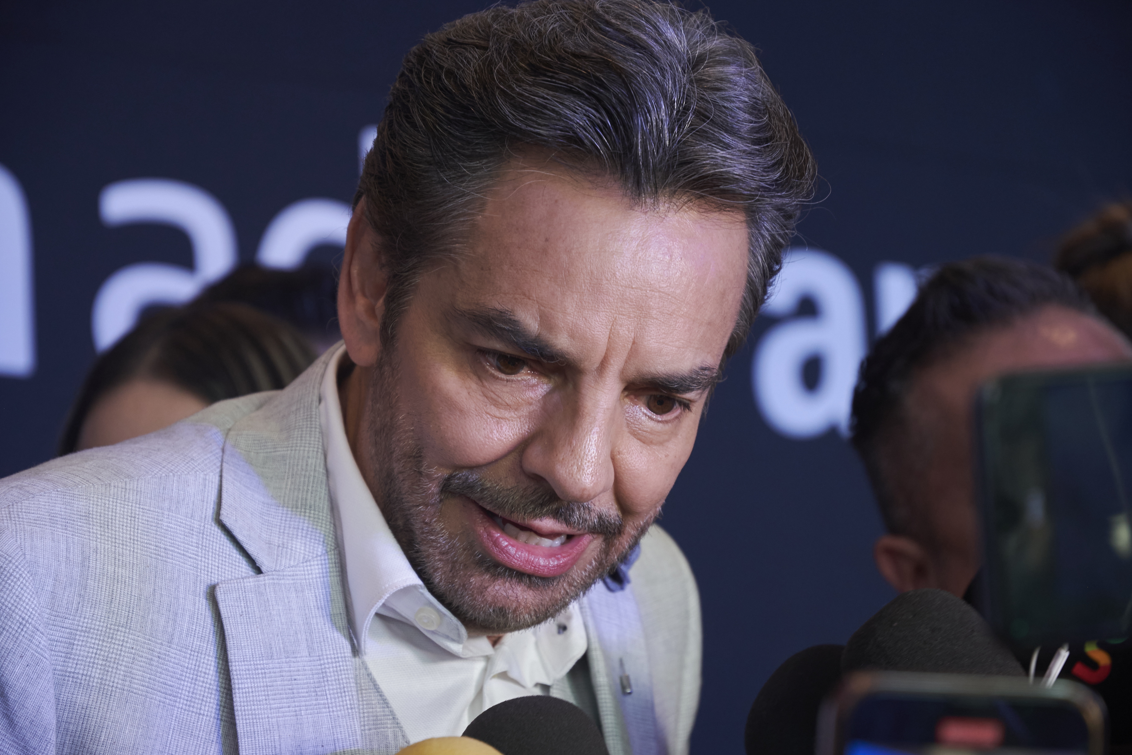 Eugenio Derbez in a suit speaking into multiple microphones at a press event