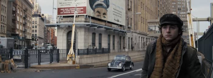 Timothée Chalamet in warm clothing and cap stands on a city street with a vintage car and a large billboard in the background