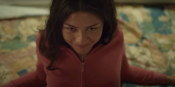 Zendaya wearing a zip-up top sitting on a patterned bedspread, looking upward