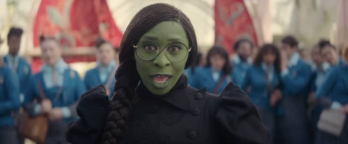 Cynthia Erivo with green face, glasses, and braided hair wearing a detailed jacket. Group of people in blue uniforms stands in the blurred background