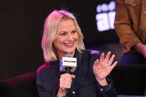 Amy Poehler smiling while speaking into a microphone at a SiriusXM event, wearing a button-down shirt and seated on a stage
