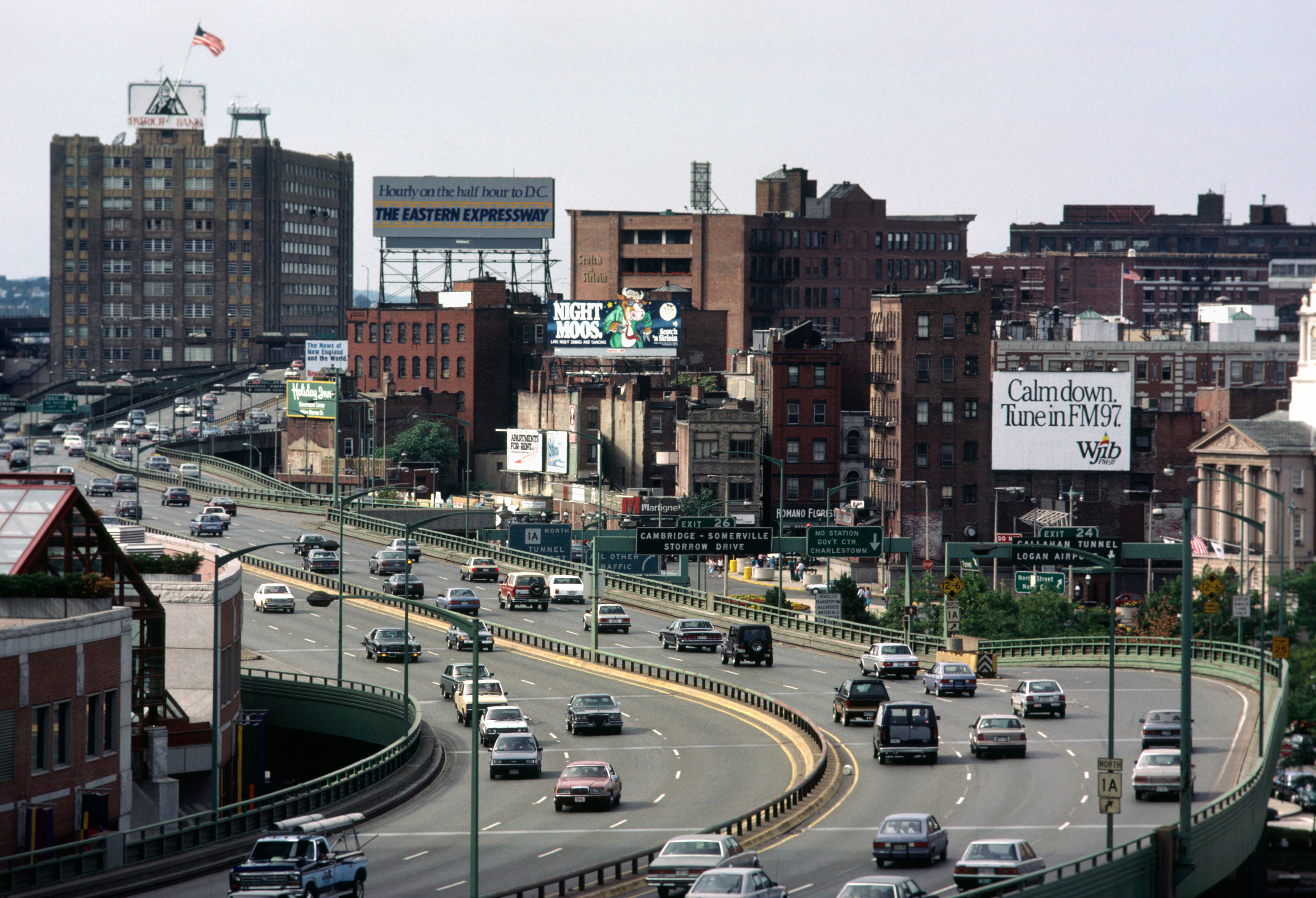 City scene with vintage cars on a busy highway, bordered by brick buildings and billboards. Urban atmosphere with a mix of traffic and architecture