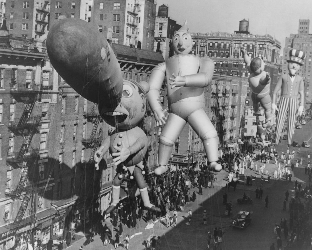 Inflatable cartoon character balloons float above a crowded street during a parade in a historic urban setting