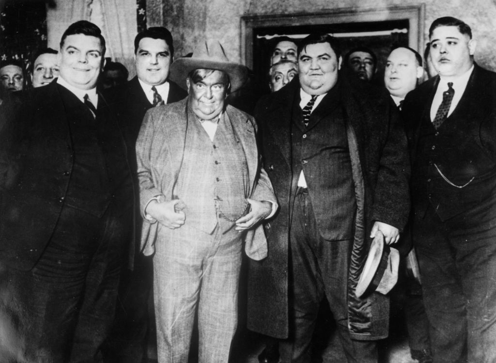 Group of well-dressed men from the early 20th century gather indoors. Some smile, and one wears a hat. The setting suggests a formal event