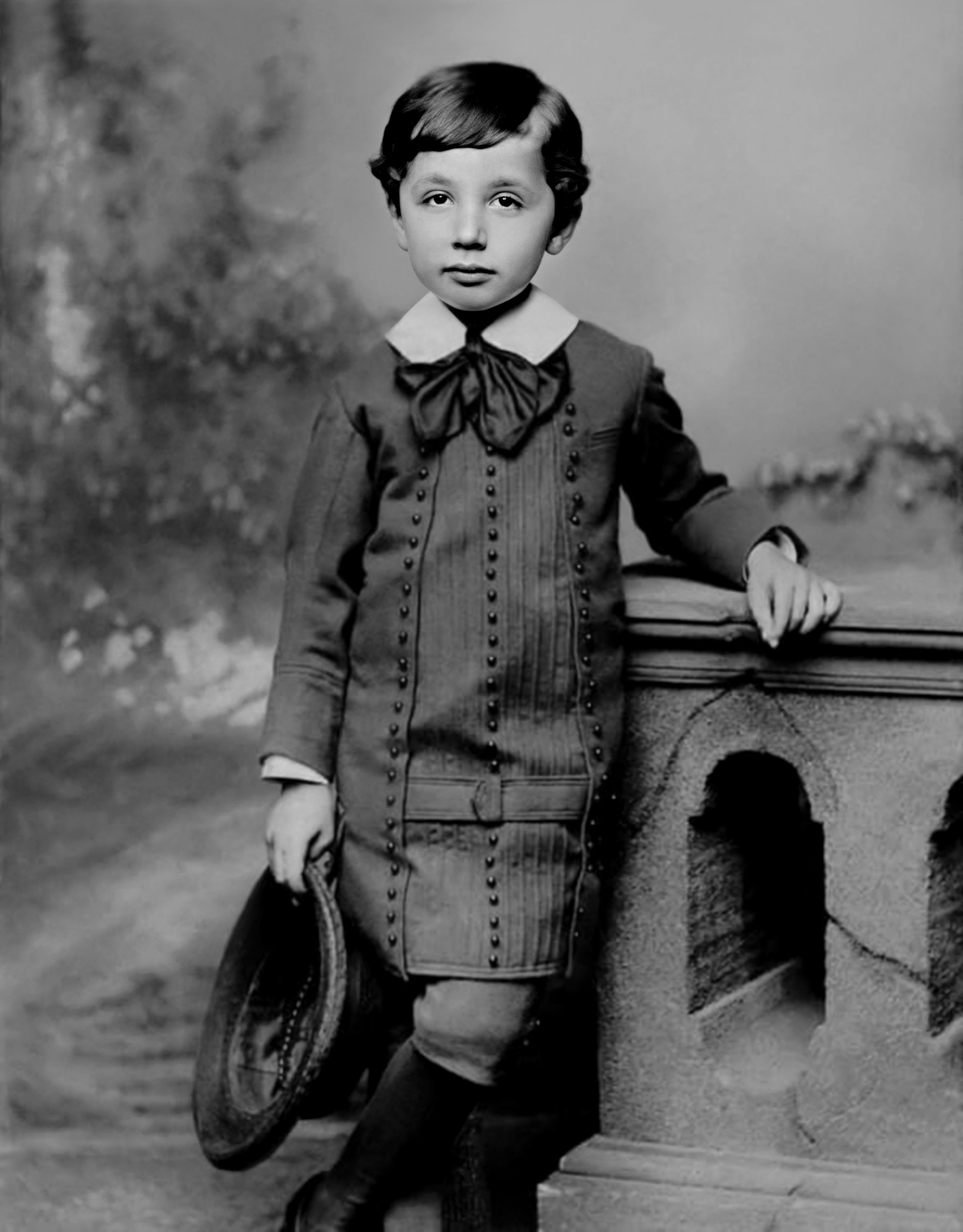 A young child stands in a formal outfit holding a hat, posing for a vintage portrait