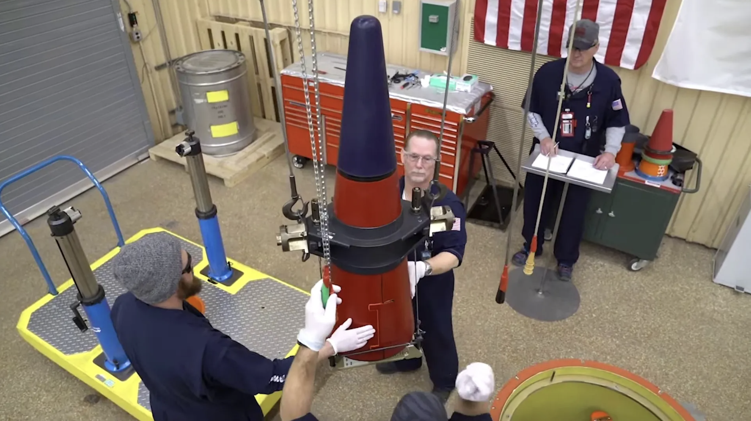 People in a workshop are assembling a large red and black device using a lifting mechanism. An American flag is in the background