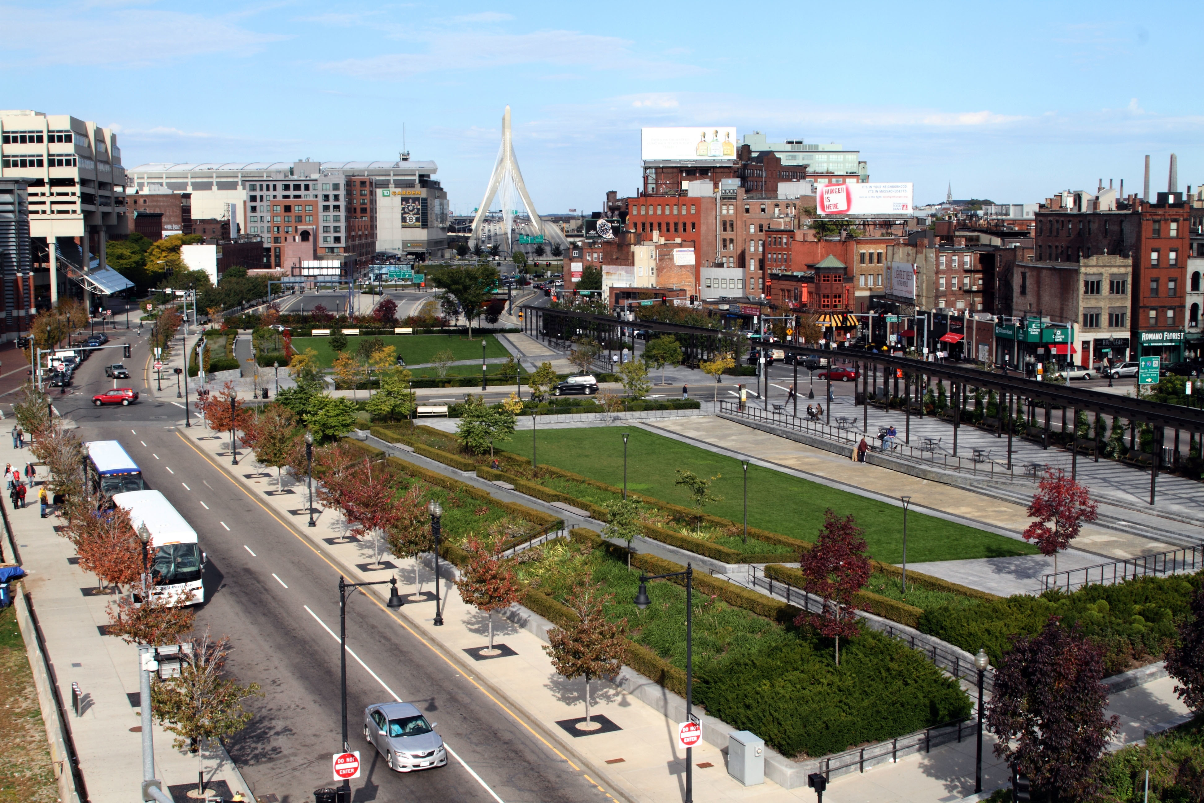 City park with green lawns, trees, and pathways, surrounded by urban buildings and a distant bridge under a clear sky