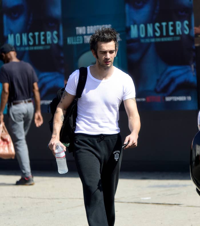 Matty Healy in a white T-shirt and black sweatpants holding a water bottle, walking outside with movie posters in the background