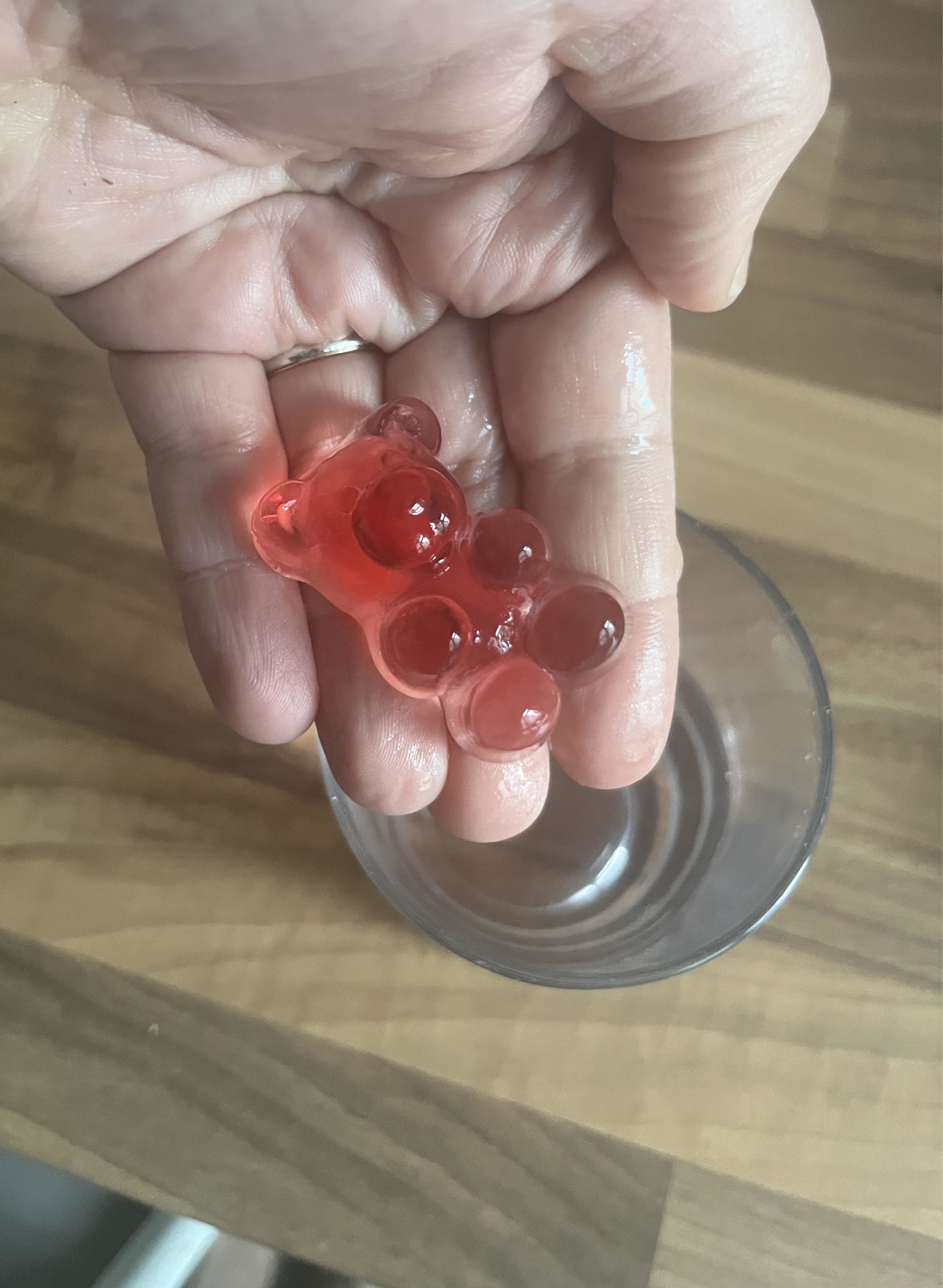 A hand holds a gummy bear formed by merging several smaller gummy bears with a glass below