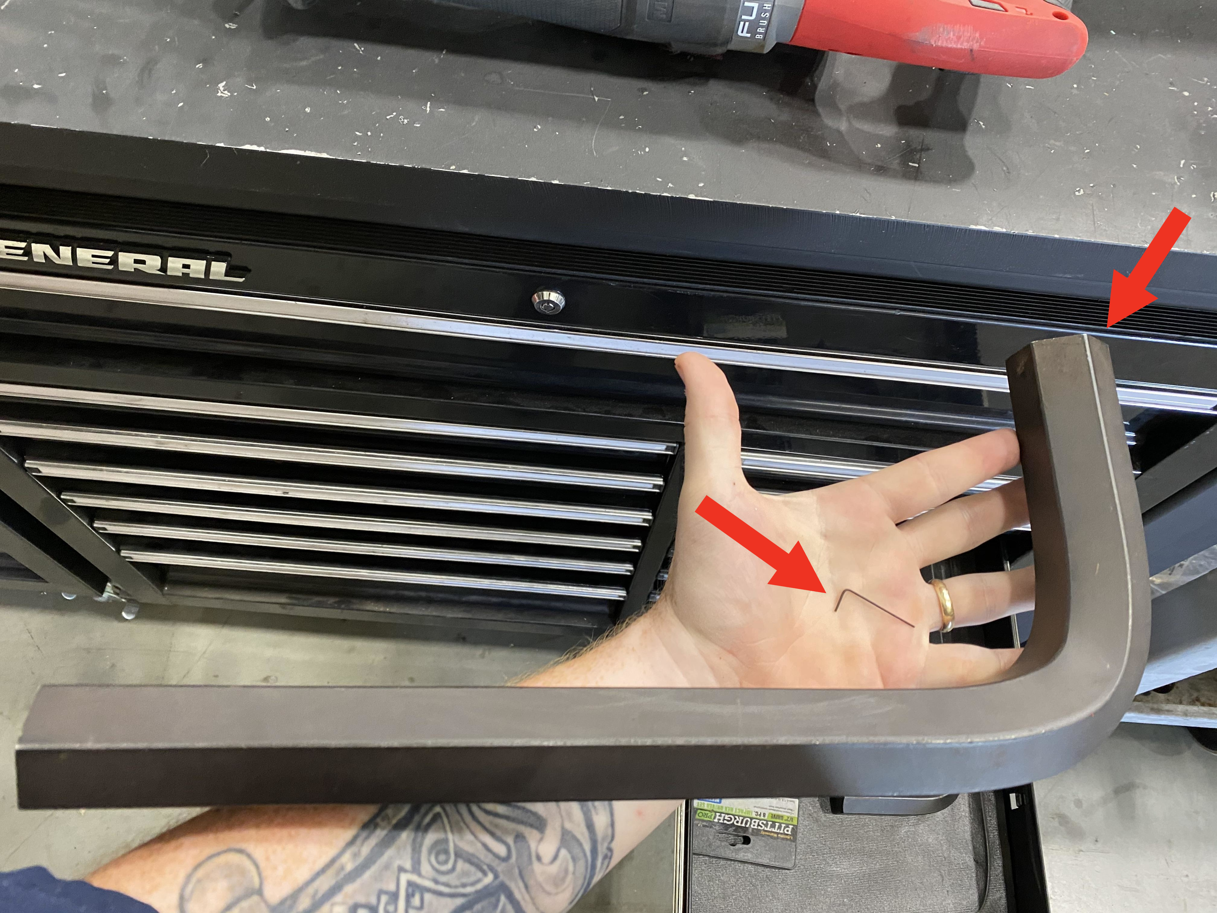 A hand holding a large L-shaped hex key near a tool chest, showcasing the size of the tool