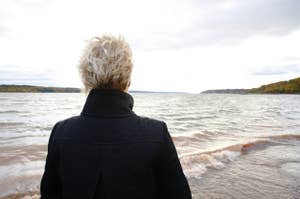 Person with short blond hair stands facing a large body of water, wearing a dark jacket. The scene appears serene and contemplative