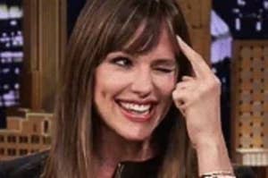 Person smiling on a talk show set, pointing to their forehead, wearing a black outfit with long sleeves and layered bracelets