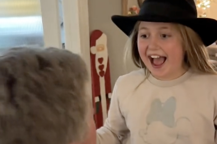 帽子をかぶった若い女の子が笑顔で部屋に入り、前にいる人物に驚いている様子。