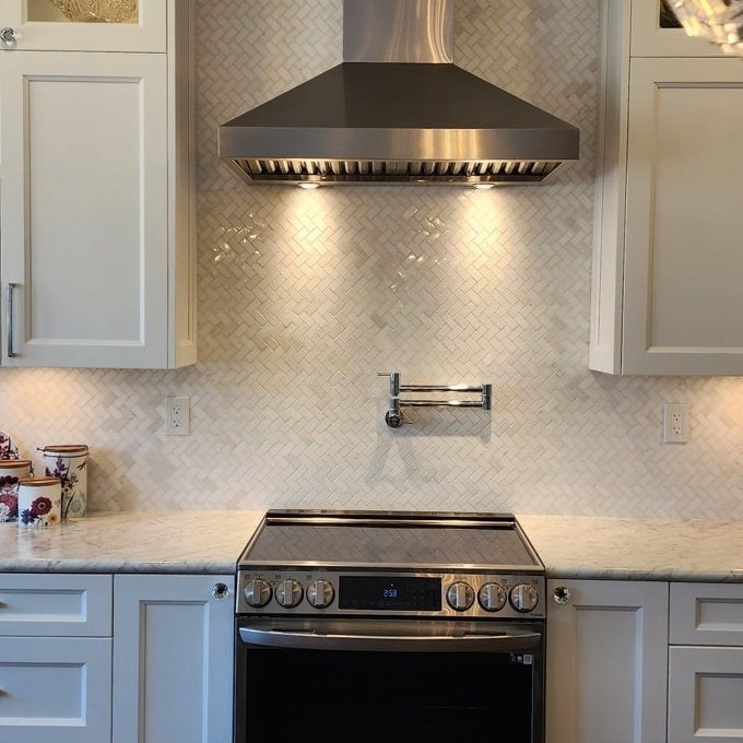 Modern kitchen with stainless steel hood, herringbone tile backsplash, and marble countertop featuring a stove and pull-down faucet