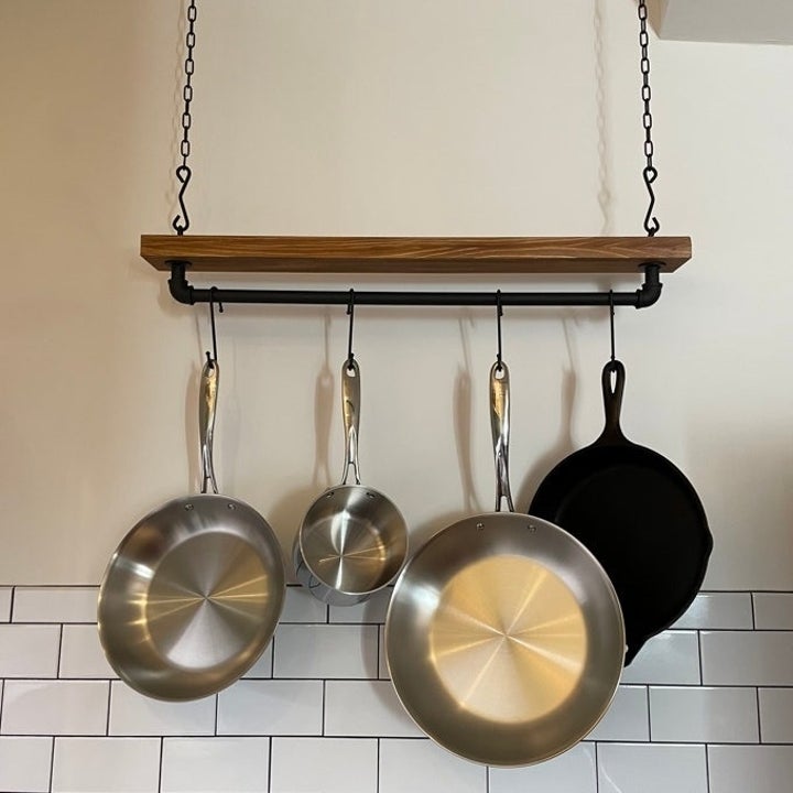 Hanging cookware set on a wall-mounted rack above a tiled backsplash, featuring stainless steel pans and a cast iron skillet