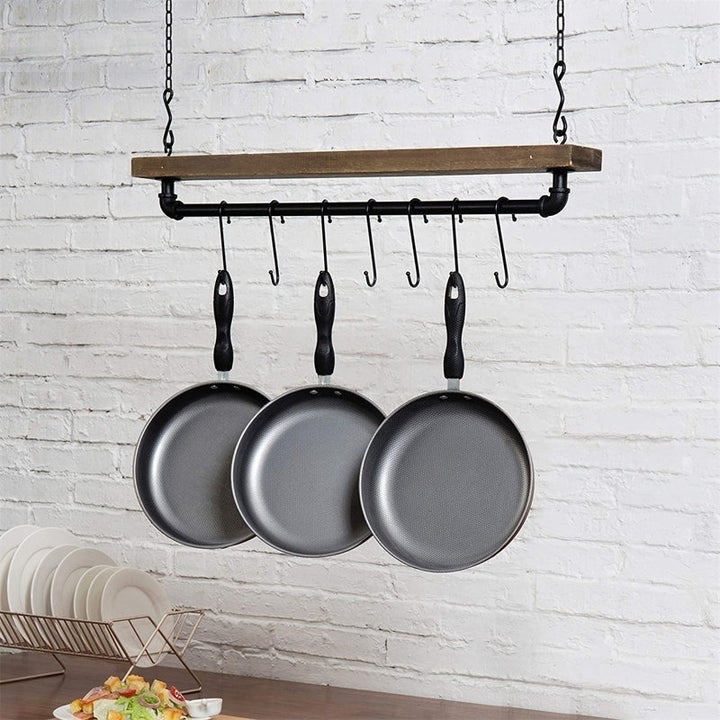 Hanging on a wooden rack with hooks are three metal frying pans, above a kitchen counter with a dish rack holding white plates