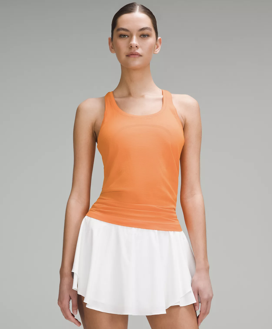 Model wearing an orange tank top and white tennis skirt in a minimalist setting