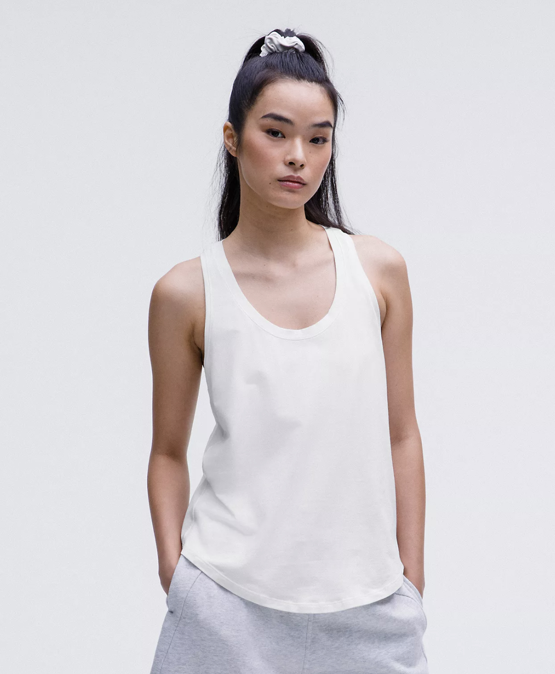 model in a sleeveless casual white tank and sweatpants, standing with hands in pockets. Hair is tied up with a scrunchie