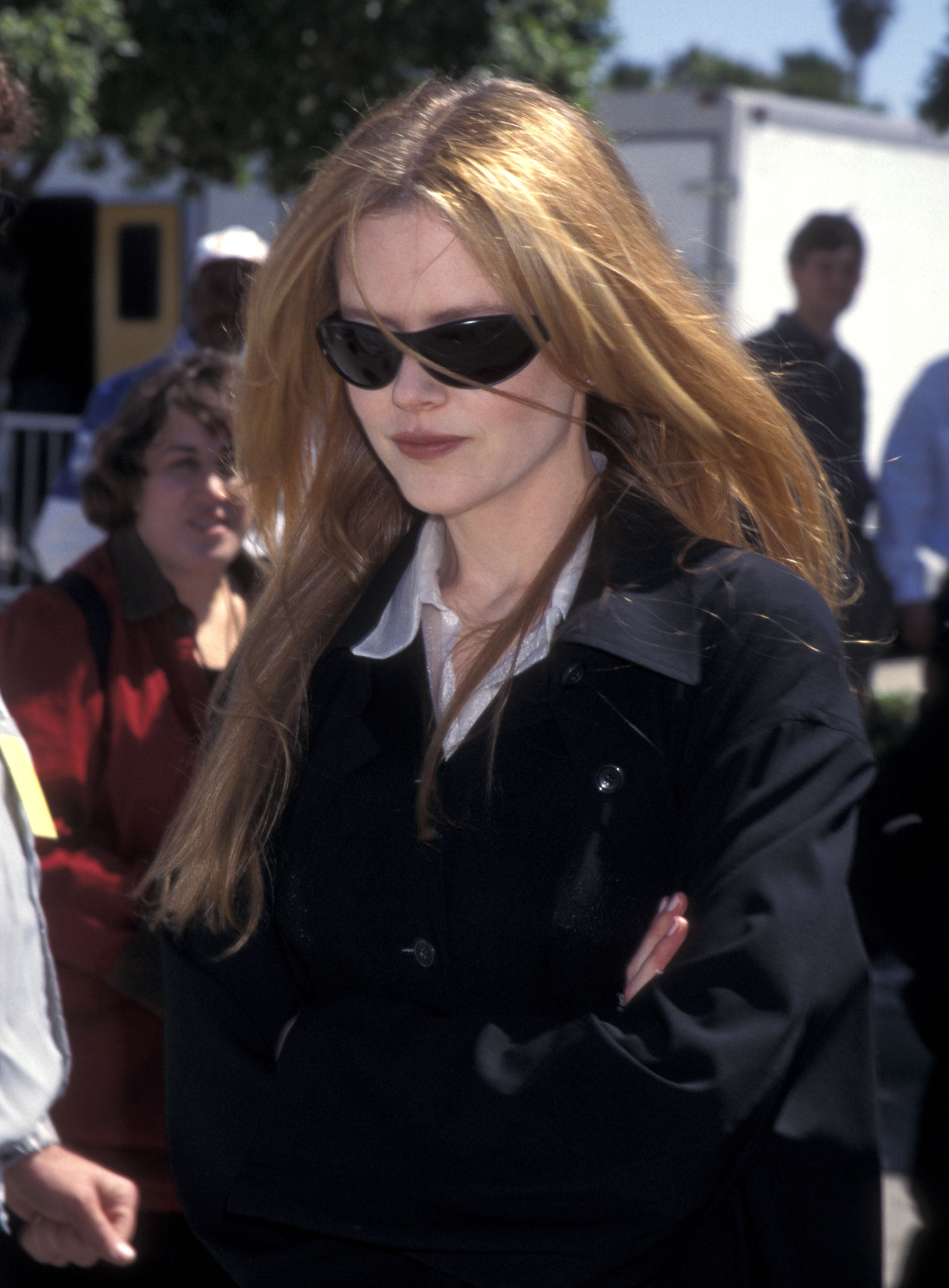 Nicole Kidman wearing sunglasses and a black coat, arms crossed, outdoors